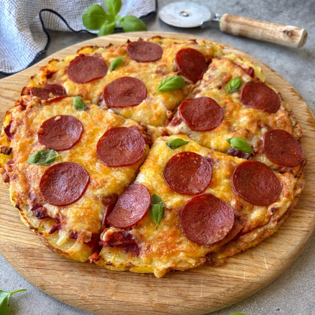 Chopping board featuring a smashed potato pizza topped with pepperoni. In the background is a pizza slicer, a checked tea towel, and fresh basil.