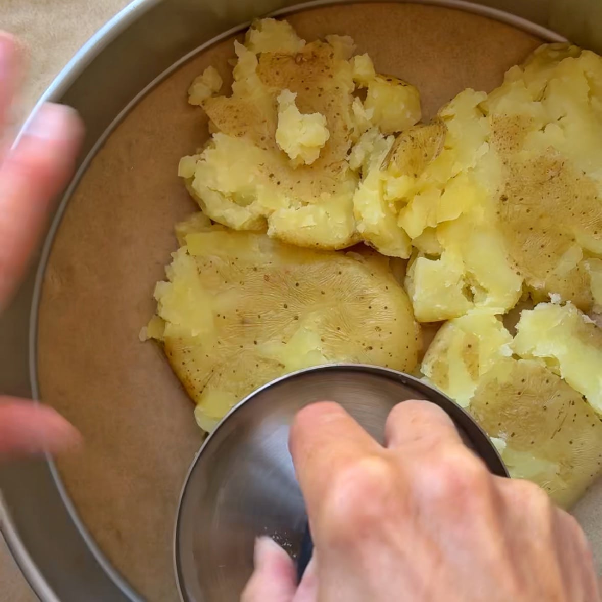A hand smashes cooked potatoes inside a round cake pan lined with parchment.