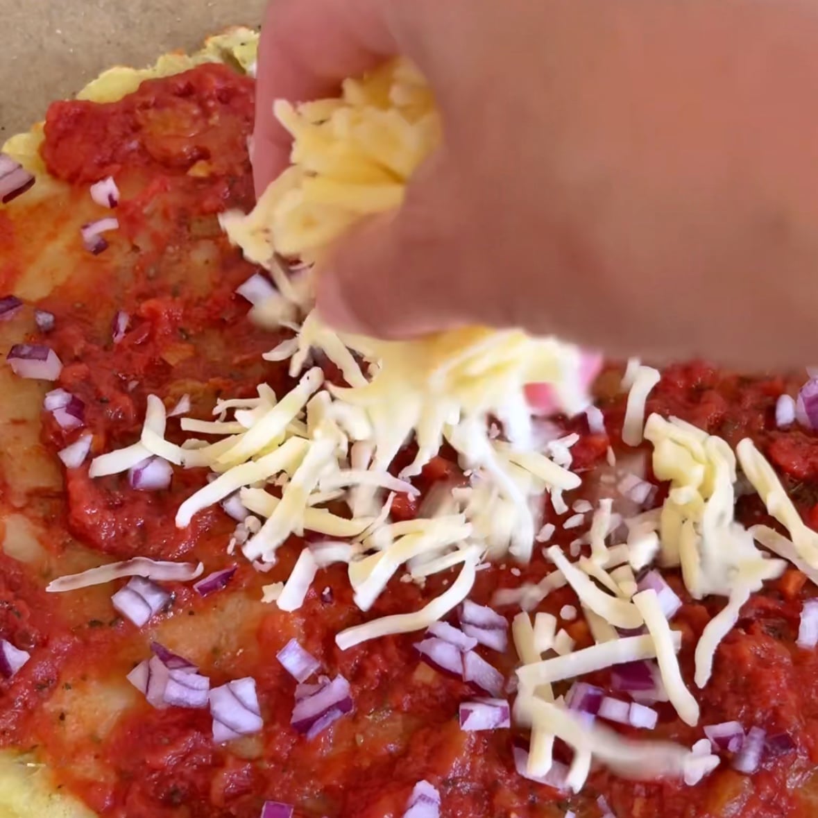 A hand sprinkles mozzarella on top of a smashed potato pizza base topped with tomato sauce and diced red onions.