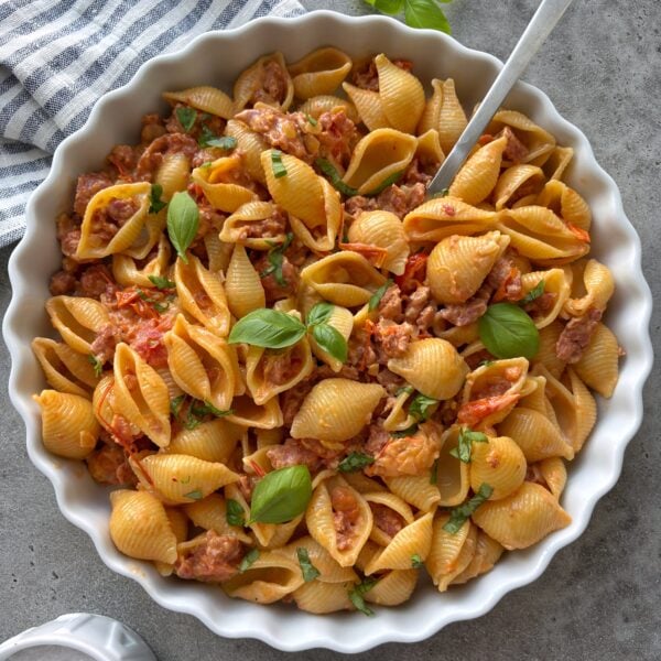 A bowl of freshly made Spicy Italian Sausage Pasta with a striped tea towel and fresh basil in the background.