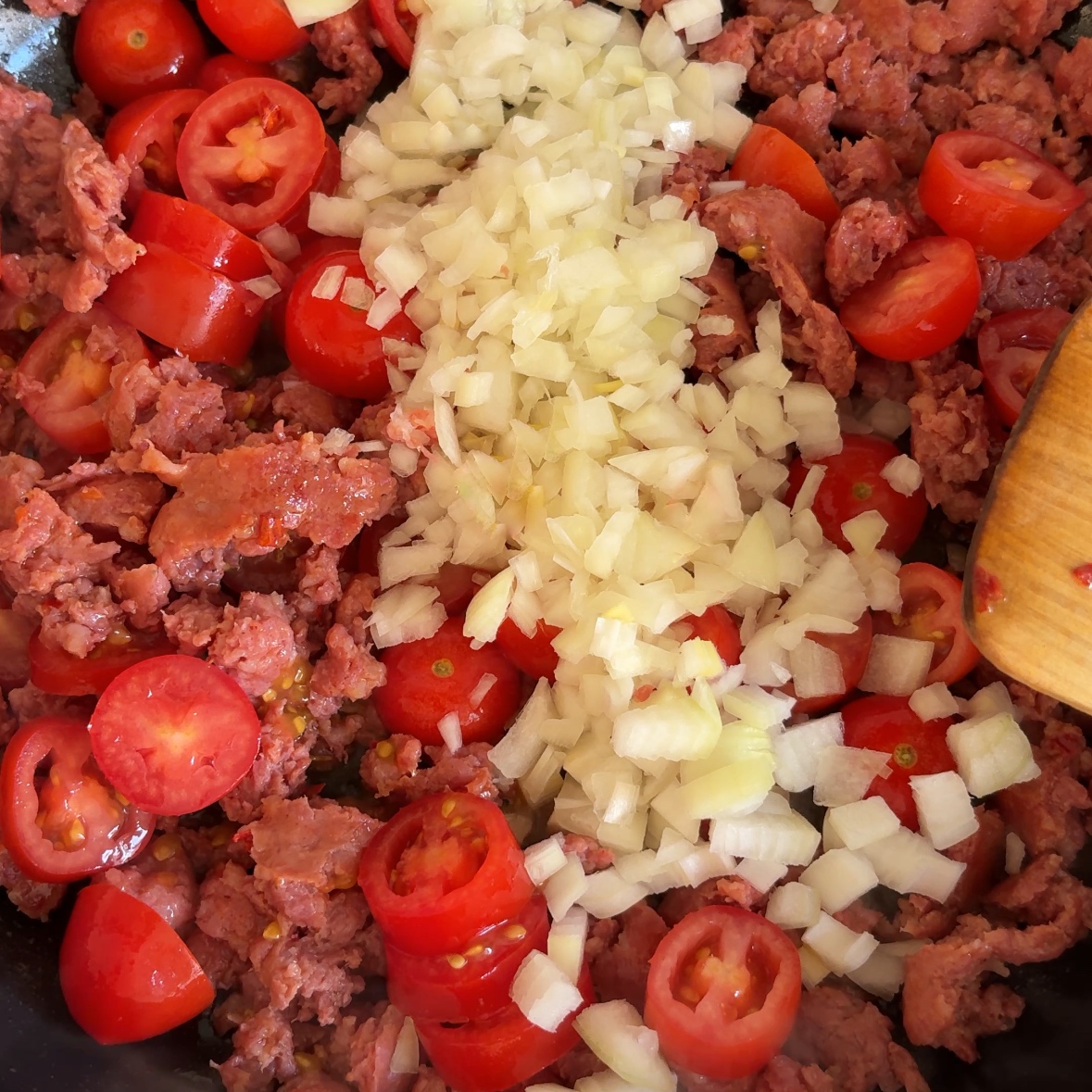 A closeup of chopped onions and tomatoes being added to spicy Italian sausage cooking in a large pan.
