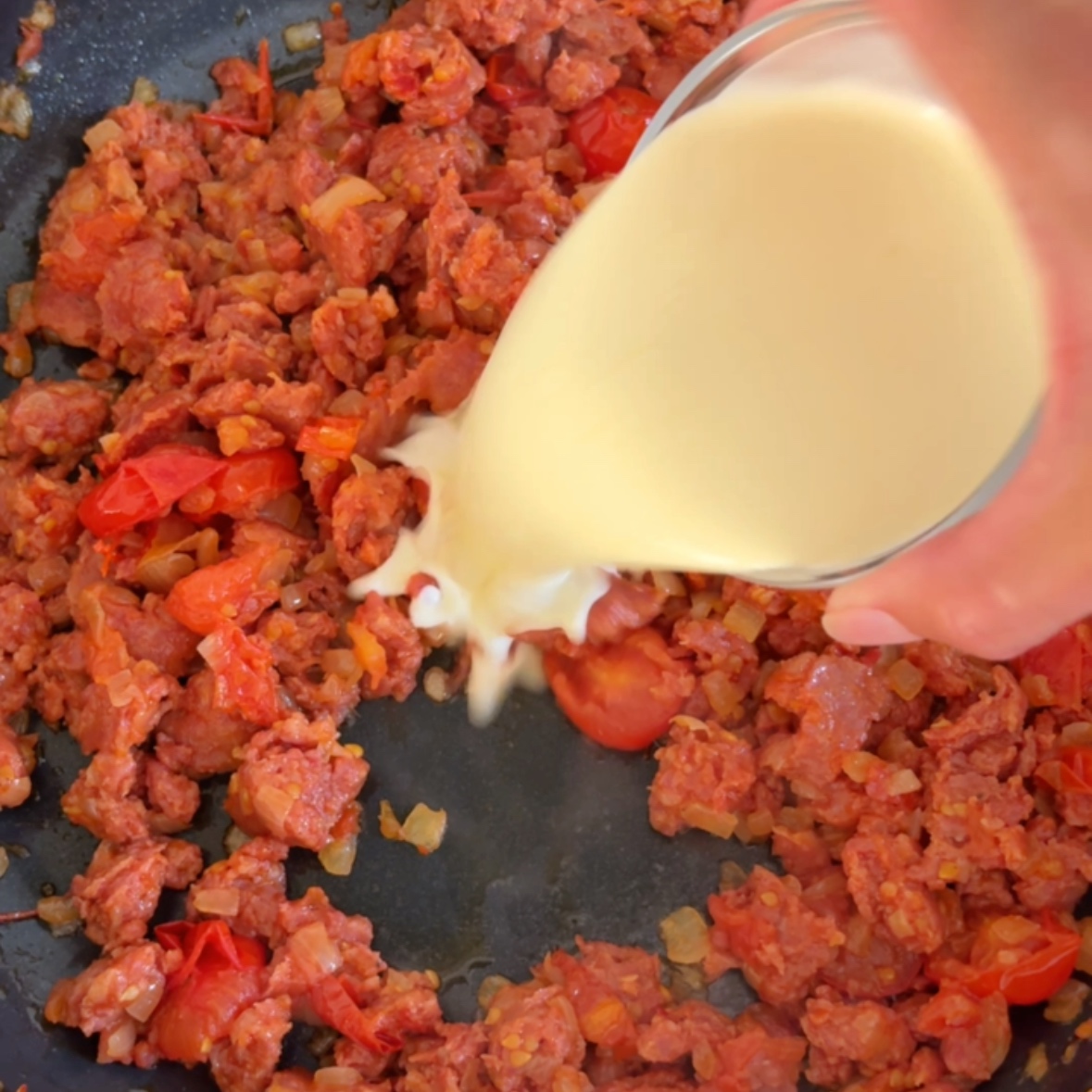 A hand pours heavy cream into a pan with spicy Italian sausage, tomatoes, and onions.