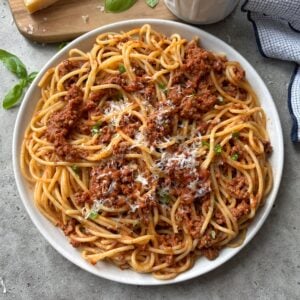 Beef bolognese topped with chopped basil and Parmesan. In the background is a block of cheese on a chopping board, fresh basil leaves, a tub of salt, and a blue checked tea towel.