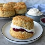 A closeup of a buttermilk scone topped with whipped cream and jam. In the background are more scones resting on a baking rack.