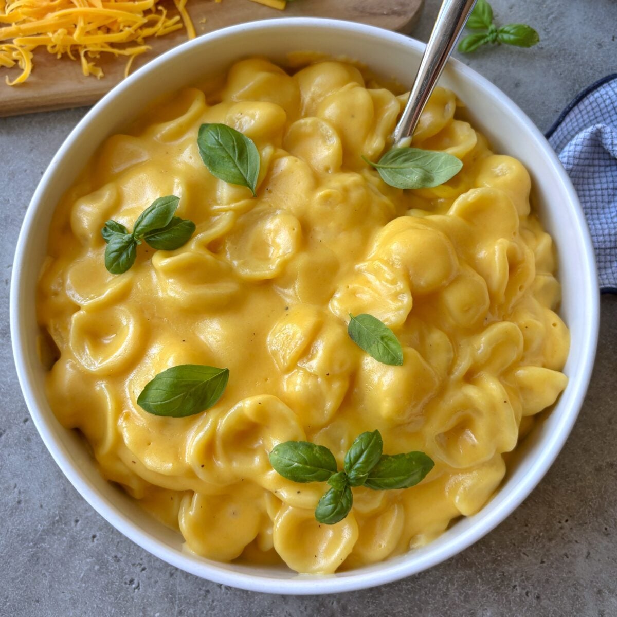 The top view of a bowl of Cheddar Mac and Cheese with shredded cheddar on a chopping board and a checked tea towel in the background.