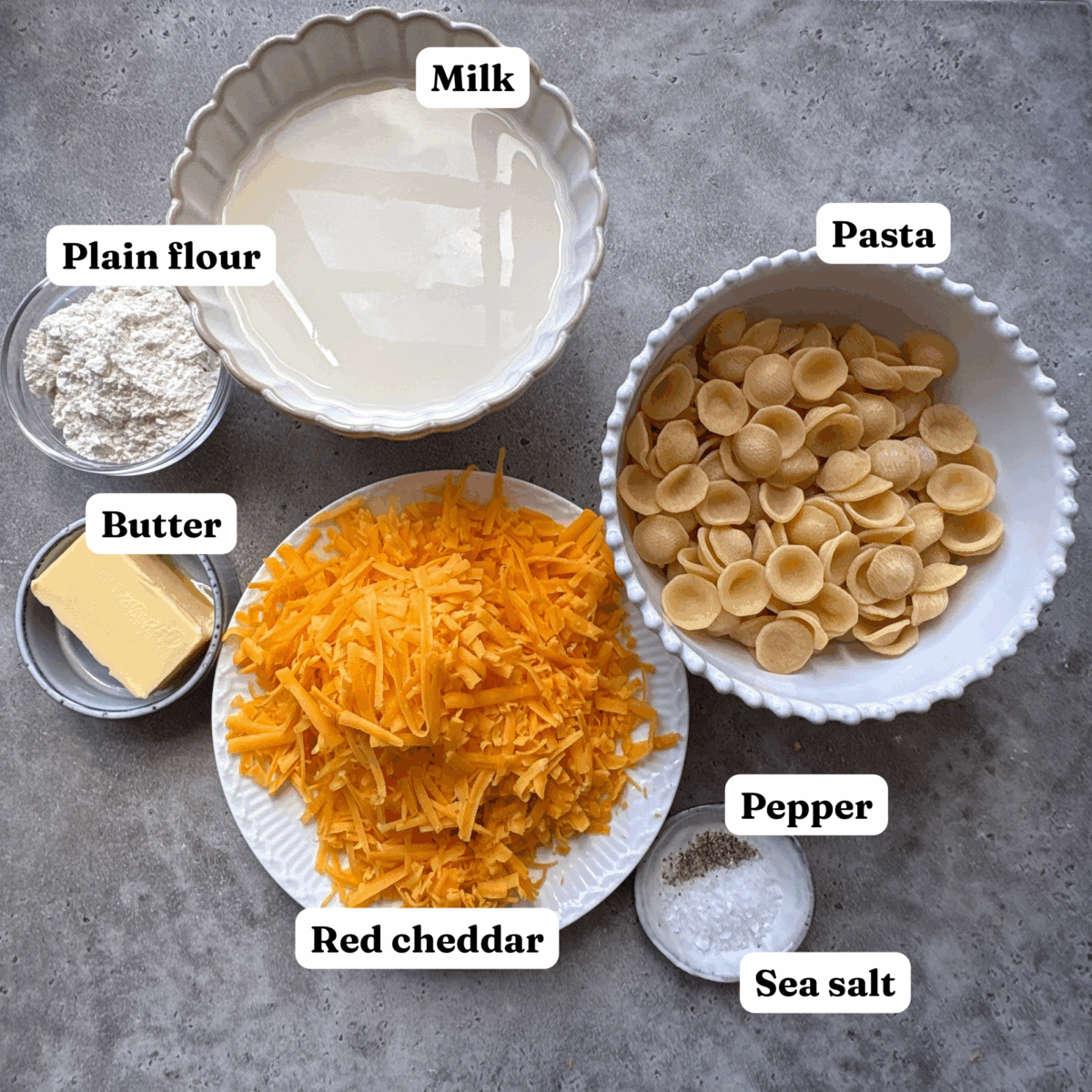 Ingredients needed for Cheddar Mac and Cheese with bowls of milk, pasta, cheddar, butter, flour, sea and and pepper.