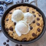 Chocolate Chip skillet cookie topped with three scoops of vanilla ice cream. In the background are more chocolate chips and a blue striped tea towel.