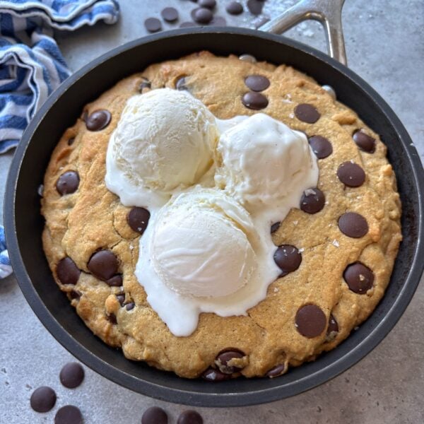 Chocolate Chip skillet cookie topped with three scoops of vanilla ice cream. In the background are more chocolate chips and a blue striped tea towel.
