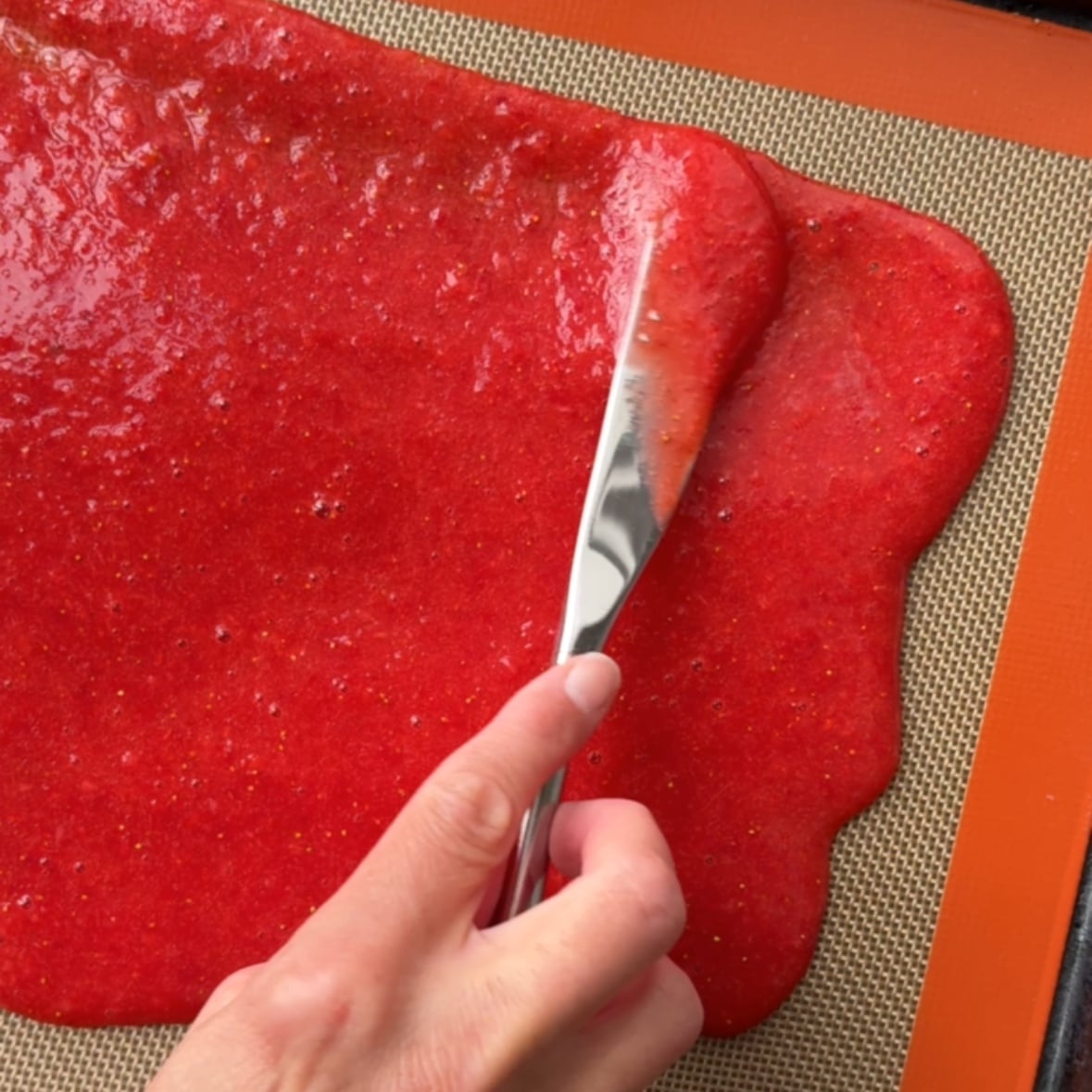 A knife spreads out pureed strawberries on a baking mat.