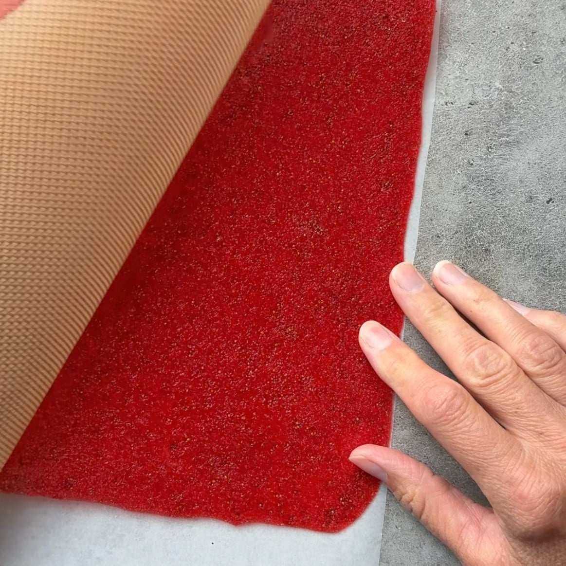 A hand separates the silicone mat from the baked strawberry fruit leather.