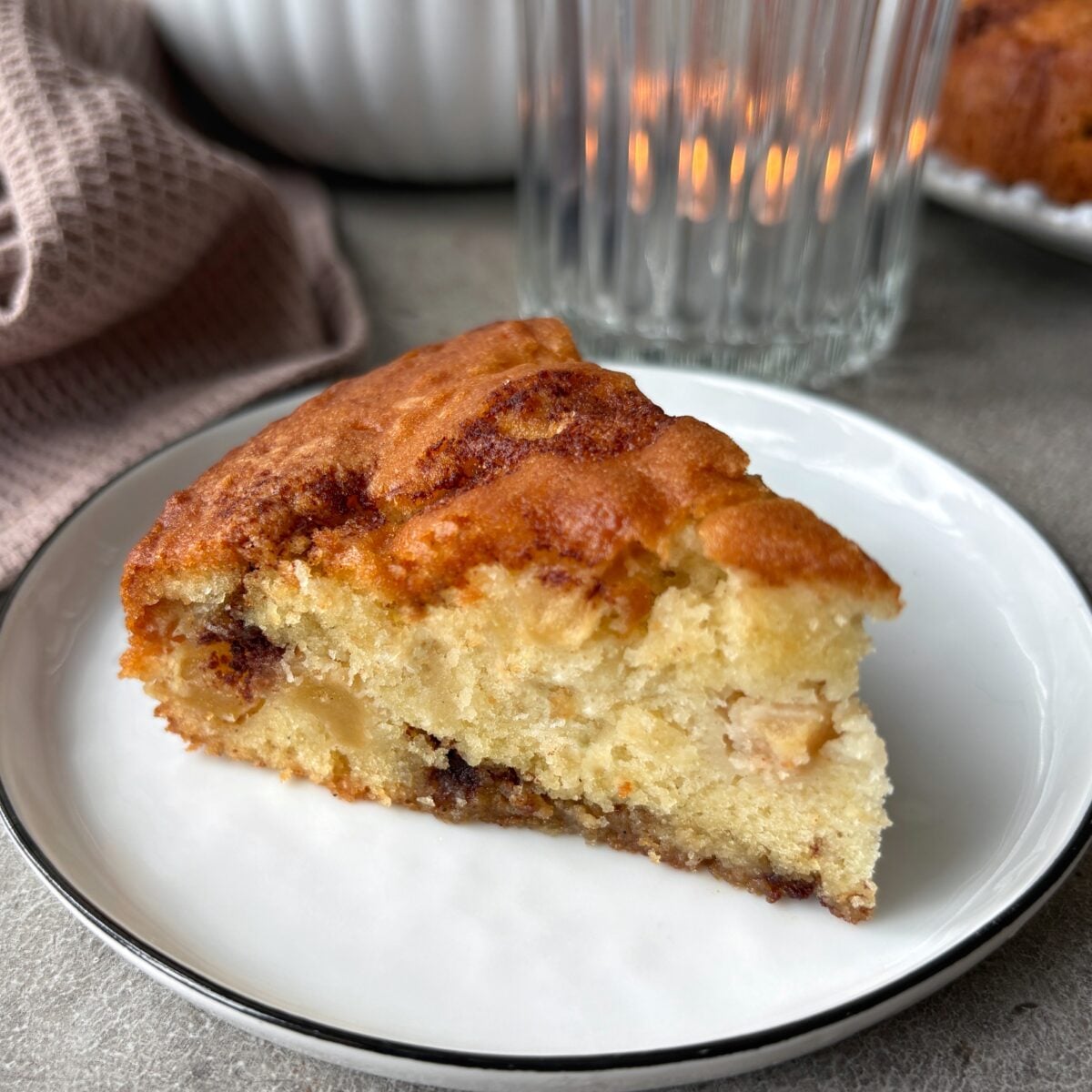 A slice of Cinnamon Apple Cake on a small white plate with a kitchen towel and a tealight in the background.