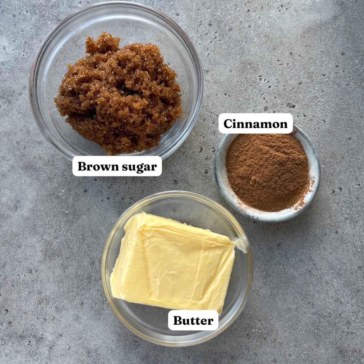 Three labeled bowls with brown sugar, cinnamon, and butter are arranged on a gray surface.