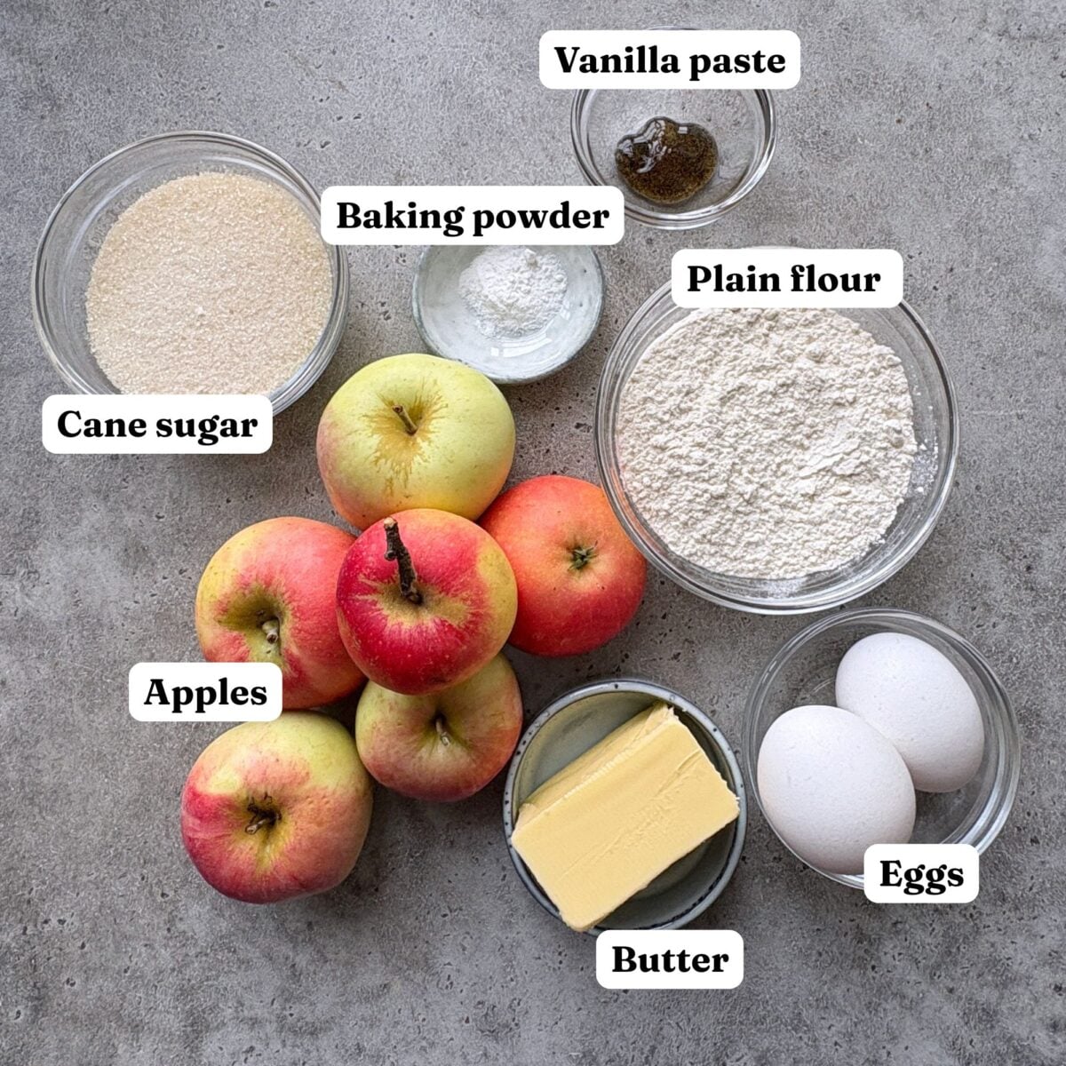 Ingredients for Cinnamon Apple Cake, featuring bowls or cane sugar, vanilla paste, eggs, butter, plain flour, baking powder, and apples.