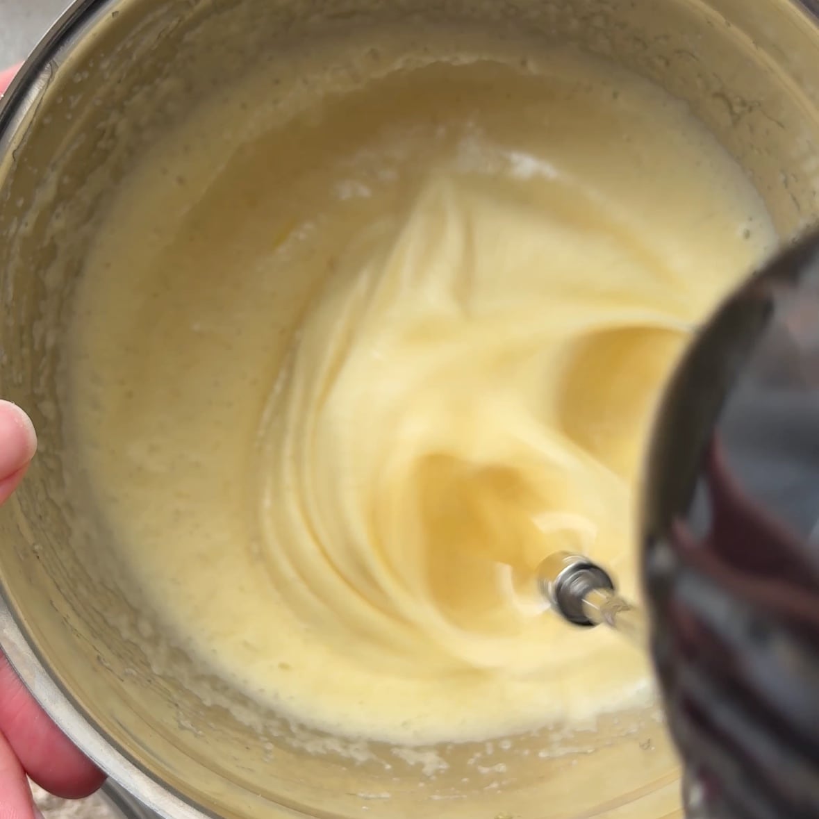 A hand holds a metal bowl while an electric mixer blends a creamy Cinnamon Apple Cake batter inside.