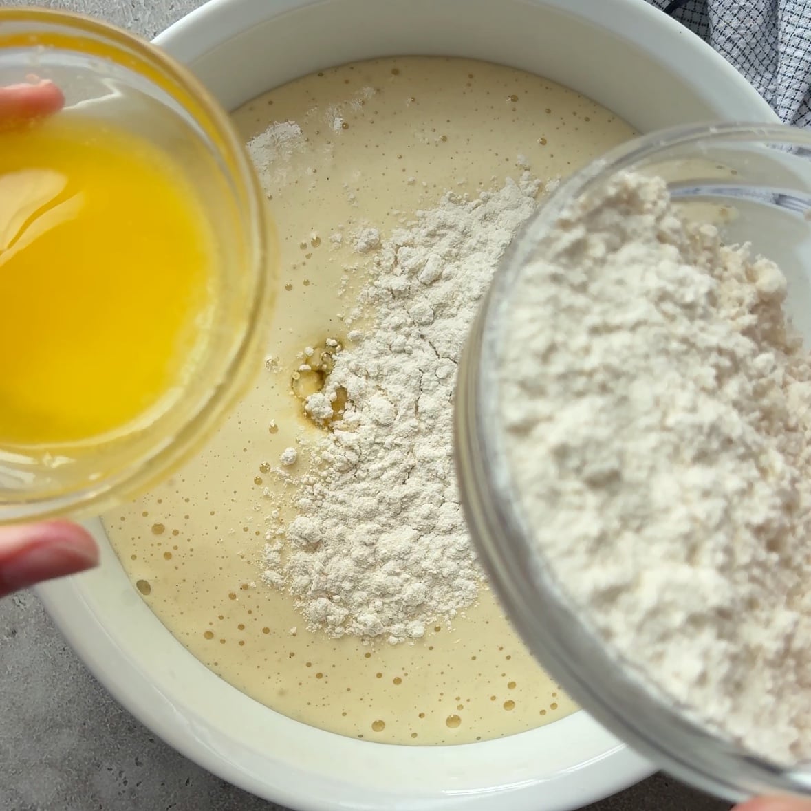 A person pours melted butter and flour into a bowl of Cinnamon Apple Cake batter on a countertop.