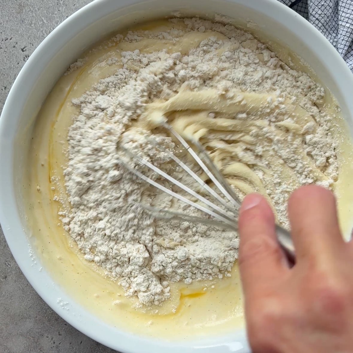 A hand uses a whisk to mix flour into a yellow batter for Cinnamon Apple Cake in a white bowl, with a kitchen towel partially visible in the corner.