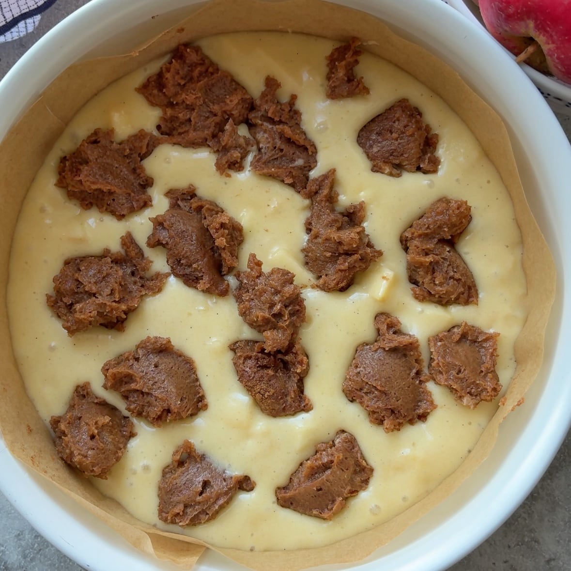A round baking dish lined with parchment paper contains pale yellow batter with uneven chunks of brown sugar filling scattered on top, ready to become a delicious Cinnamon Apple Cake.