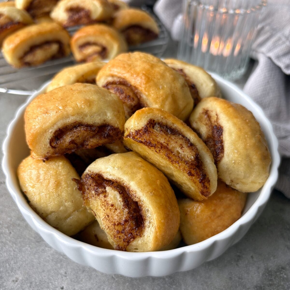 A sideview of mini cinnamon rolls in a bowl with more rolls in the background arranged on a cooling rack.