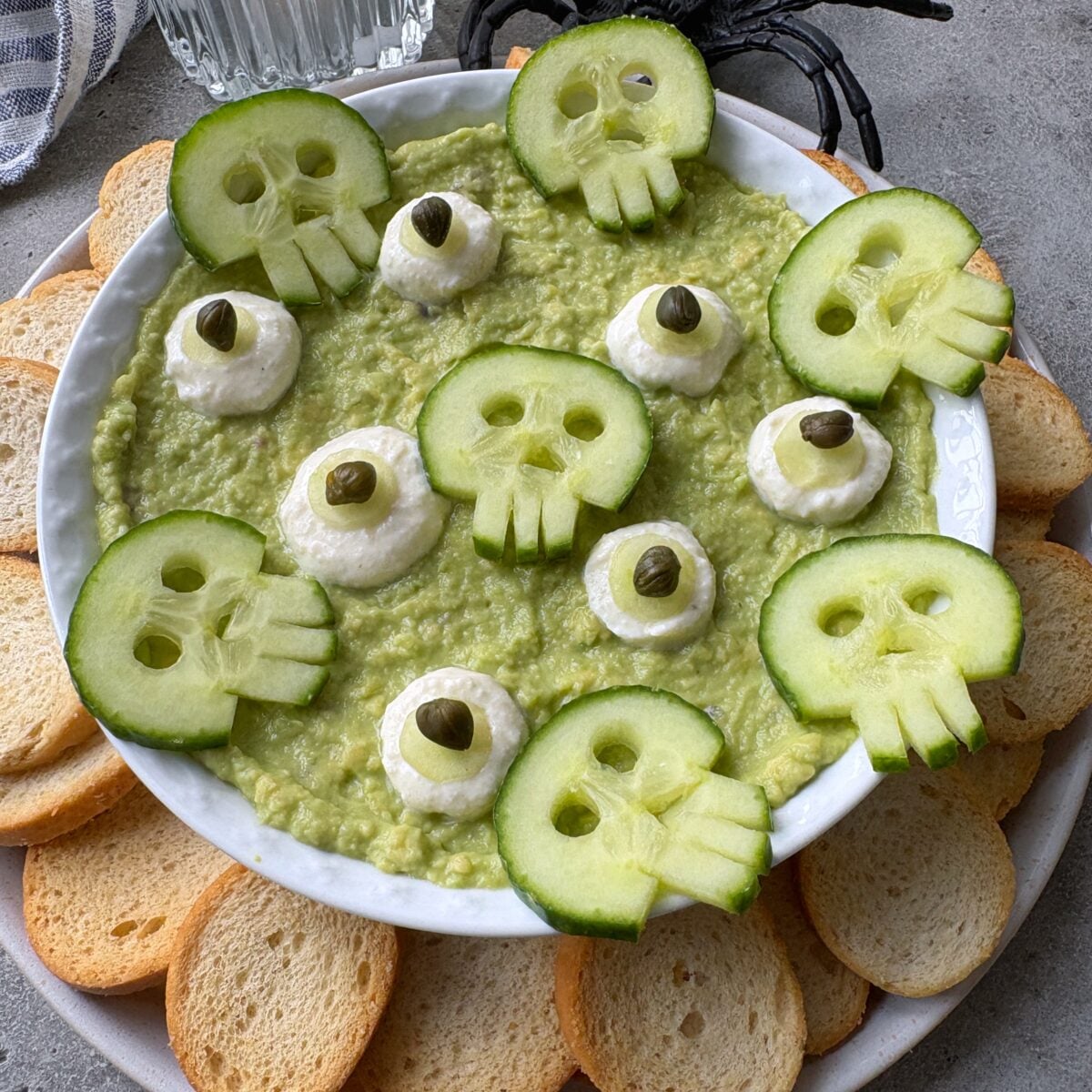 A sideview of a bowl of guacamole topped with cucumber slices cut into skull shapes, dollops of yogurt dip and capers, surrounded by round toast pieces.