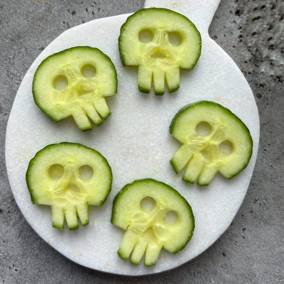 Five cucumber slices cut to resemble skull shapes are arranged on a round white cutting board placed on a gray surface.