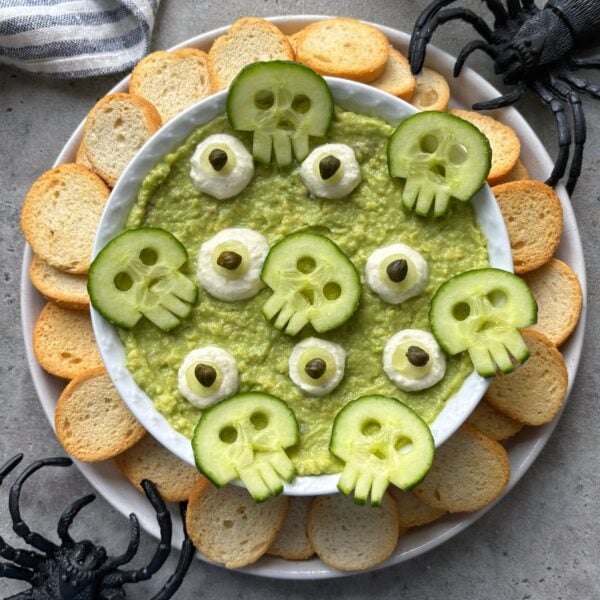 A bowl of green dip topped with cucumber skulls and surrounded by toasted bread slices, with plastic spider decorations nearby.