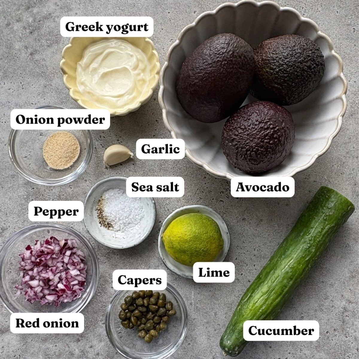 Ingredients for Halloween Guacamole are laid out including Greek yogurt, avocado, garlic, onion powder, pepper, sea salt, lime, capers, red onion, and cucumber.