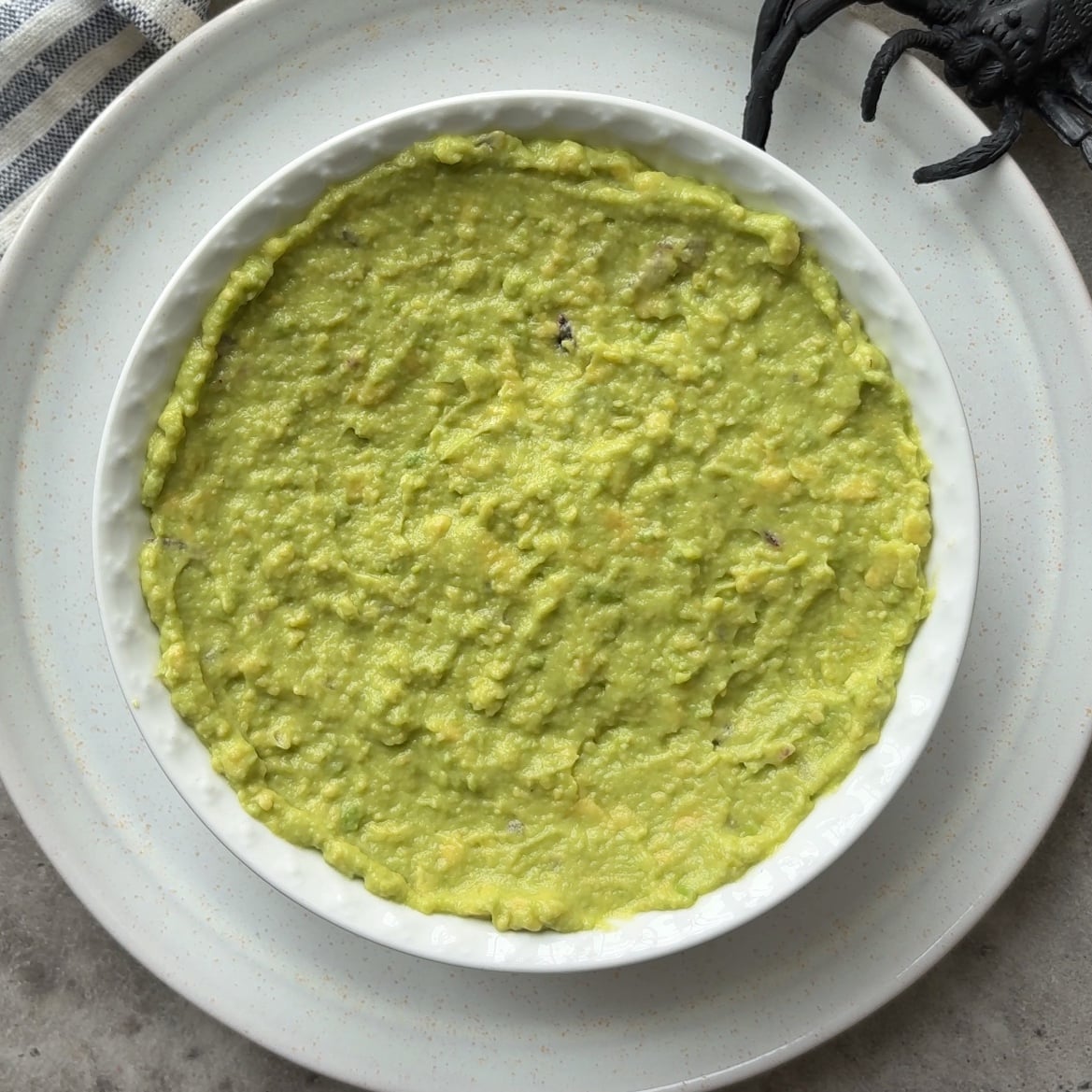 A round white dish filled with Halloween Guacamole sits on a white plate, next to a plastic black spider decoration on the top right.