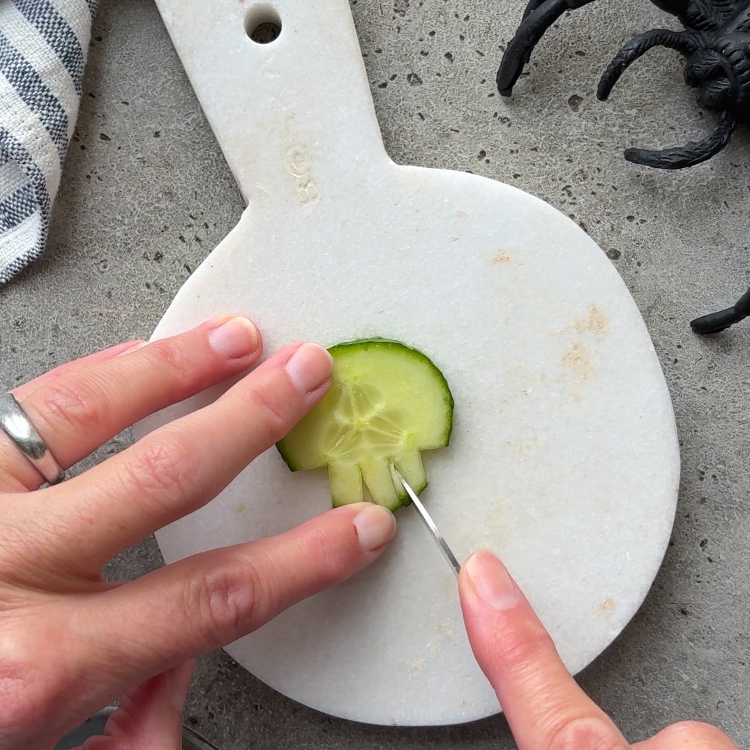 A hand carves a cucumber slice into a skull shape on a white cutting board.