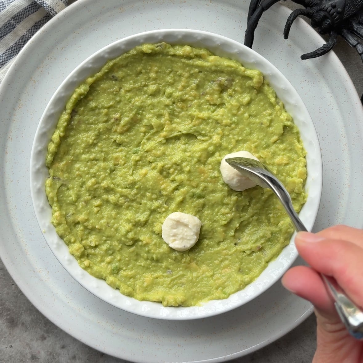 A hand uses a spoon to shape two round dollops of garlic dip into eyeballs on top of a bowl of Halloween Guacamole, with a toy spider partially visible in the background.