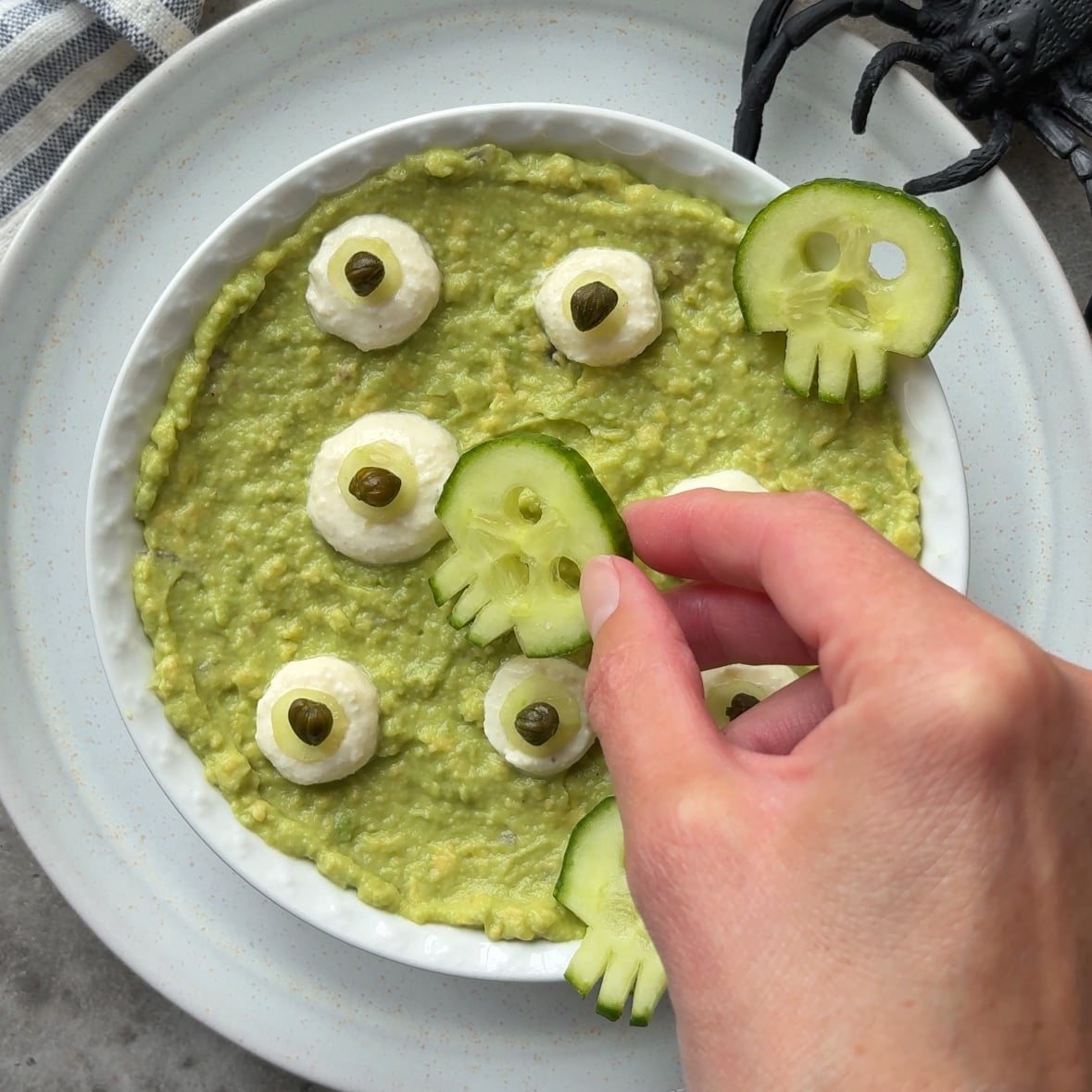 A hand places a cucumber skull on Halloween Guacamole topped with garlic dip eyeballs on a white plate with a plastic spider nearby.
