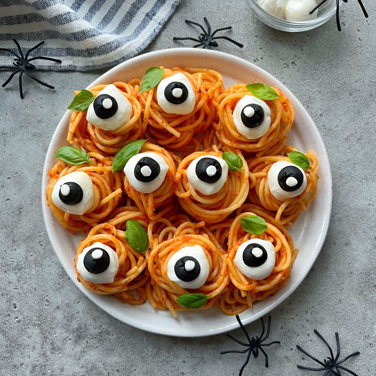 A plate of spaghetti nests topped with mozzarella balls, sliced olives, and basil leaves, arranged to look like eyeballs. Plastic spiders and a striped napkin are nearby.