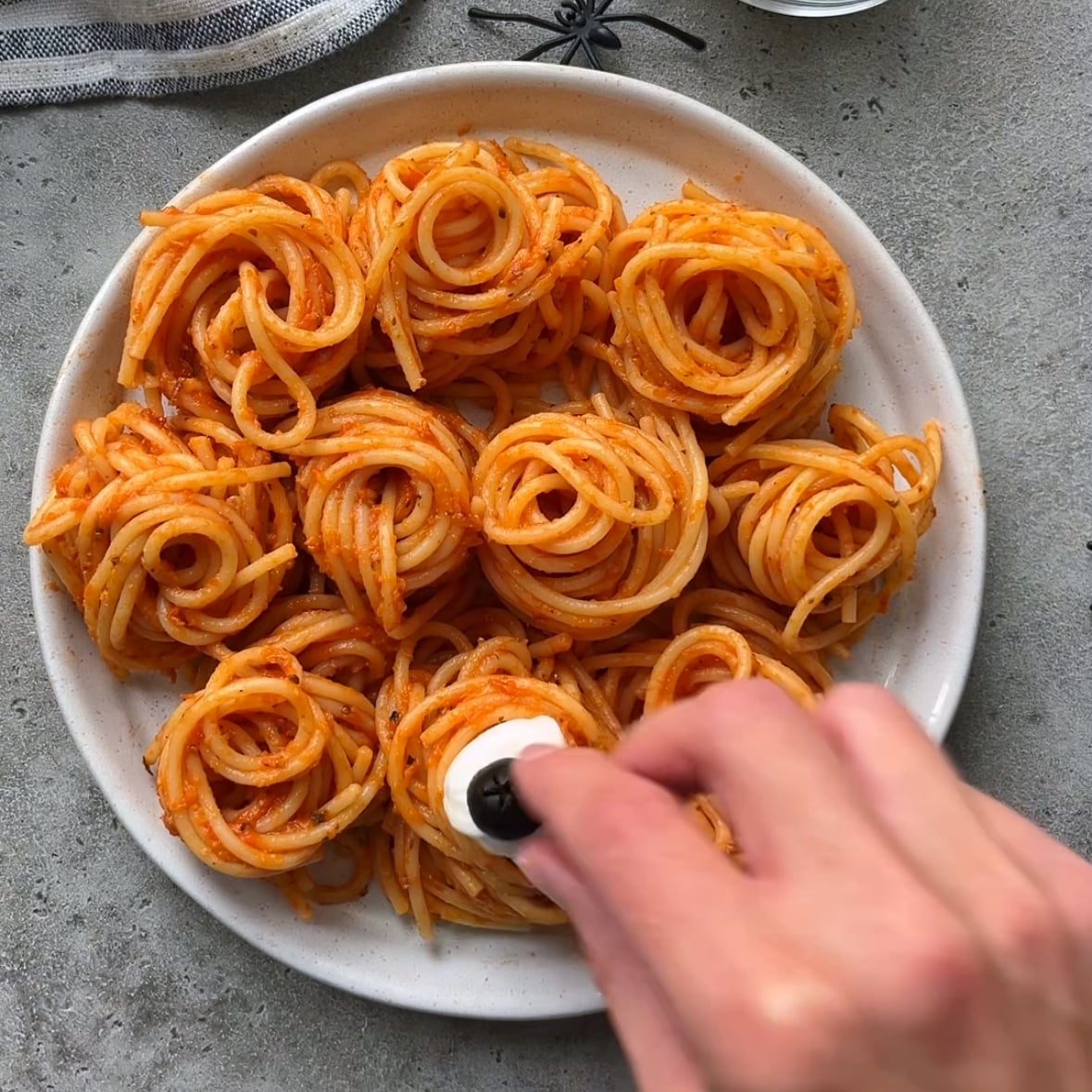 A hand tops tomato pasta nests with mozzarella and olive eyeballs.